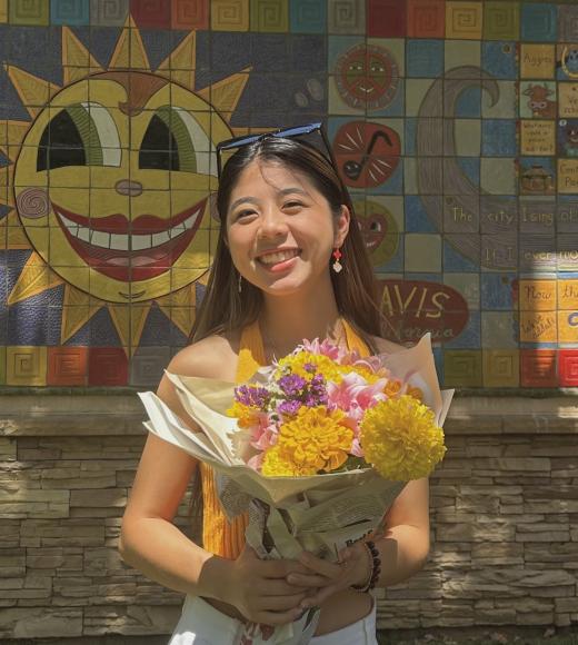profile photo of Amy wearing a white dress and holding a bouquet of yellow flowers. In the background is a colorful mural with a sun with a face and blue square tiles