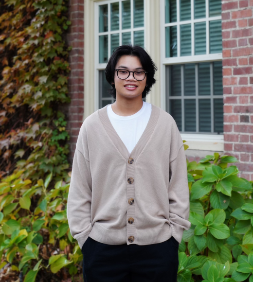 profile photo of Ayden wearing a light brown cardigan and black pants standing behind a brick building with windows