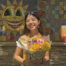 profile photo of Amy wearing a white dress and holding a bouquet of yellow flowers. In the background is a colorful mural with a sun with a face and blue square tiles
