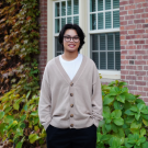 profile photo of Ayden wearing a light brown cardigan and black pants standing behind a brick building with windows